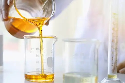 A honey-colored substance is being poured from one piece of lab glass to another piece of lab glass. Only a hand is pictured performing the action.